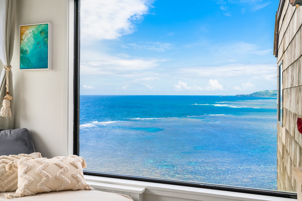 A large window reveals expansive ocean views, showcasing the vibrant blue water extending to the horizon. The shoreline and distant land form a picturesque backdrop. A cozy daybed, adorned with decorative pillows, invites relaxation while enjoying the scenic vista.