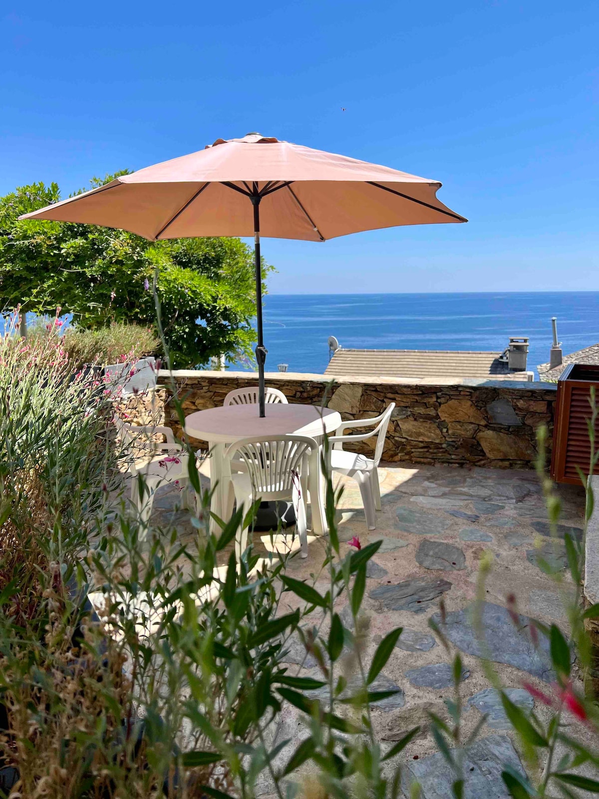 A stone patio offers a round table surrounded by white chairs, topped with a large umbrella providing shade. Lush greenery frames the scene, while the expansive view of the sea and blue sky creates a serene backdrop.