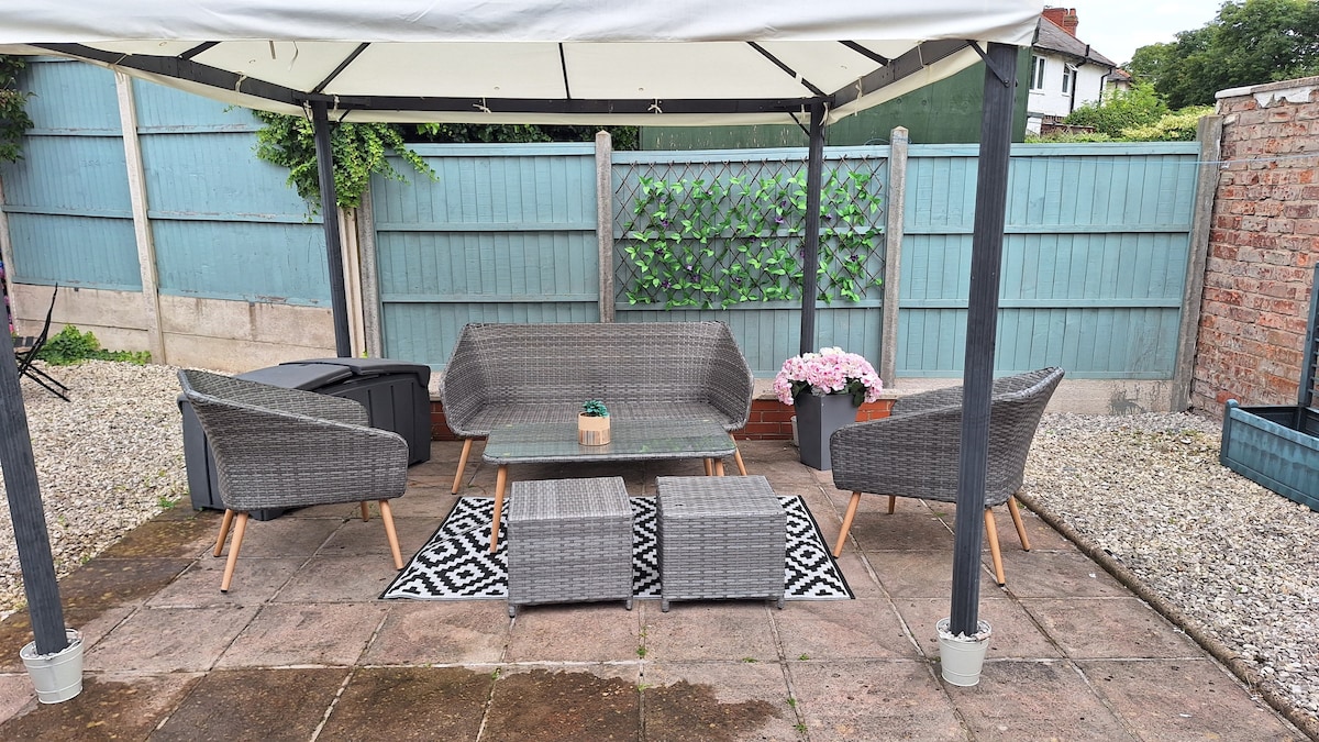 A private garden area is showcased under a gazebo, featuring a rattan patio set with a two-seater sofa, two armchairs, and a glass-topped table. Decorative planters with flowers are positioned nearby, and a patterned rug enhances the seating space. The surroundings include a gravel area and greenery.