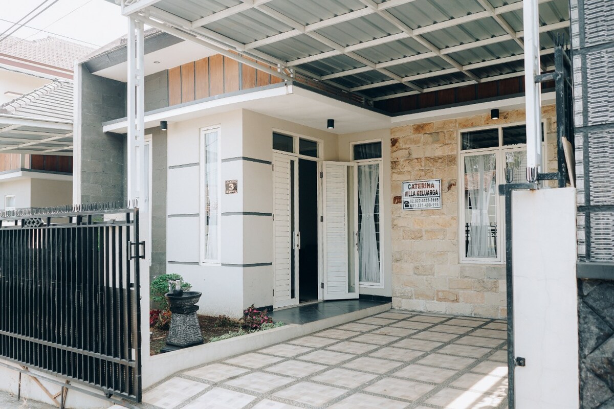 The villa features a modern facade combining light-colored walls with stone accents. A spacious entrance is framed by large windows, allowing natural light to enter. A pathway leads to the front door, bordered by landscaped areas, providing an inviting approach.