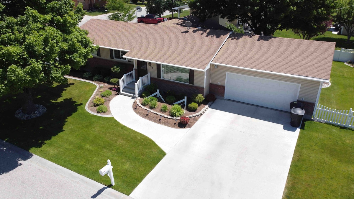 A well-maintained single-story home is set in a lush green landscape. A sloped driveway leads to the attached garage. The front yard features neatly arranged flower beds and bushes along the curved pathway, with a white fence partially visible in the background.