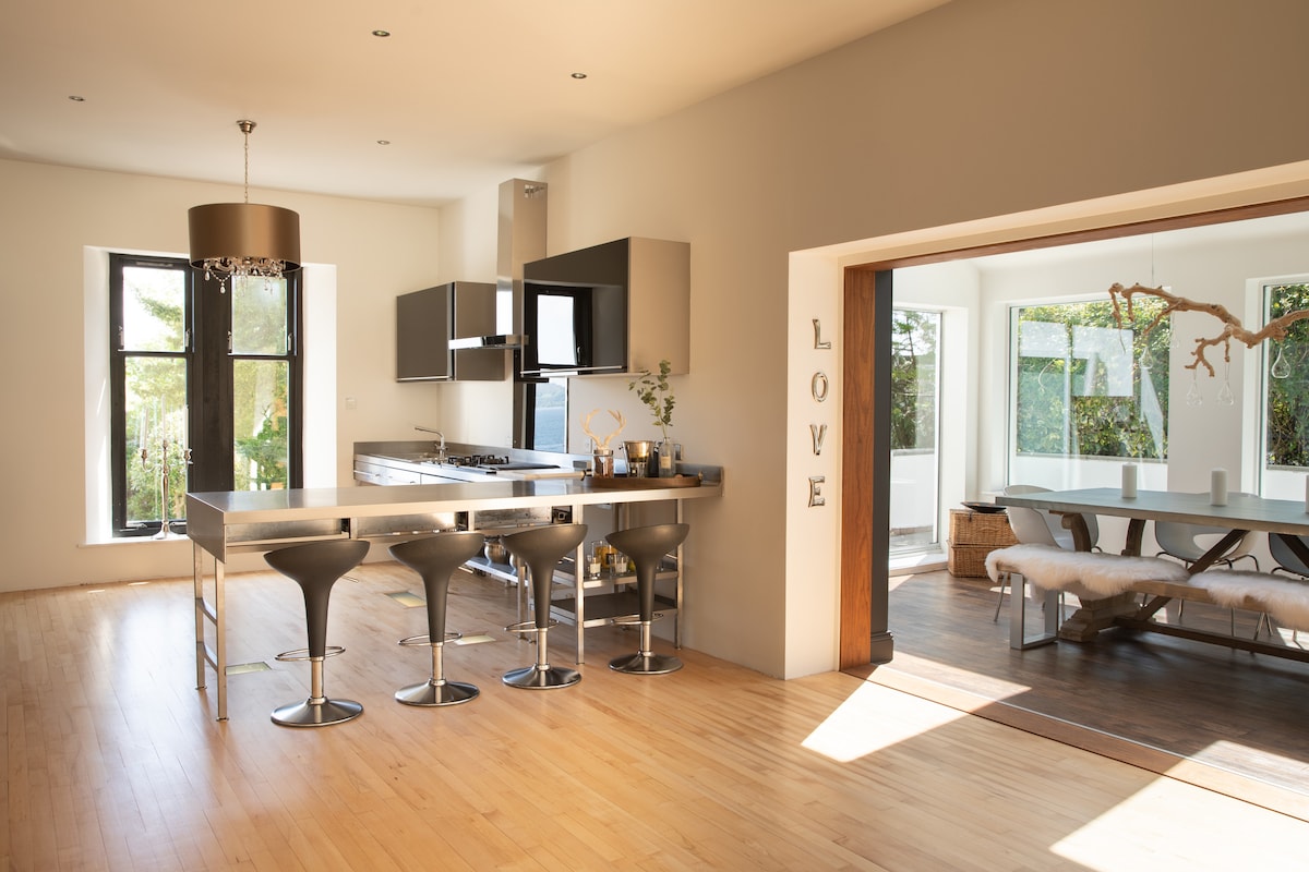 An open kitchen features a modern stainless steel countertop with seating at three high stools. Natural light fills the space, highlighting sleek cabinetry and stainless steel appliances. The adjacent dining area is visible, showcasing a long wooden table and a mix of natural and contemporary design elements.
