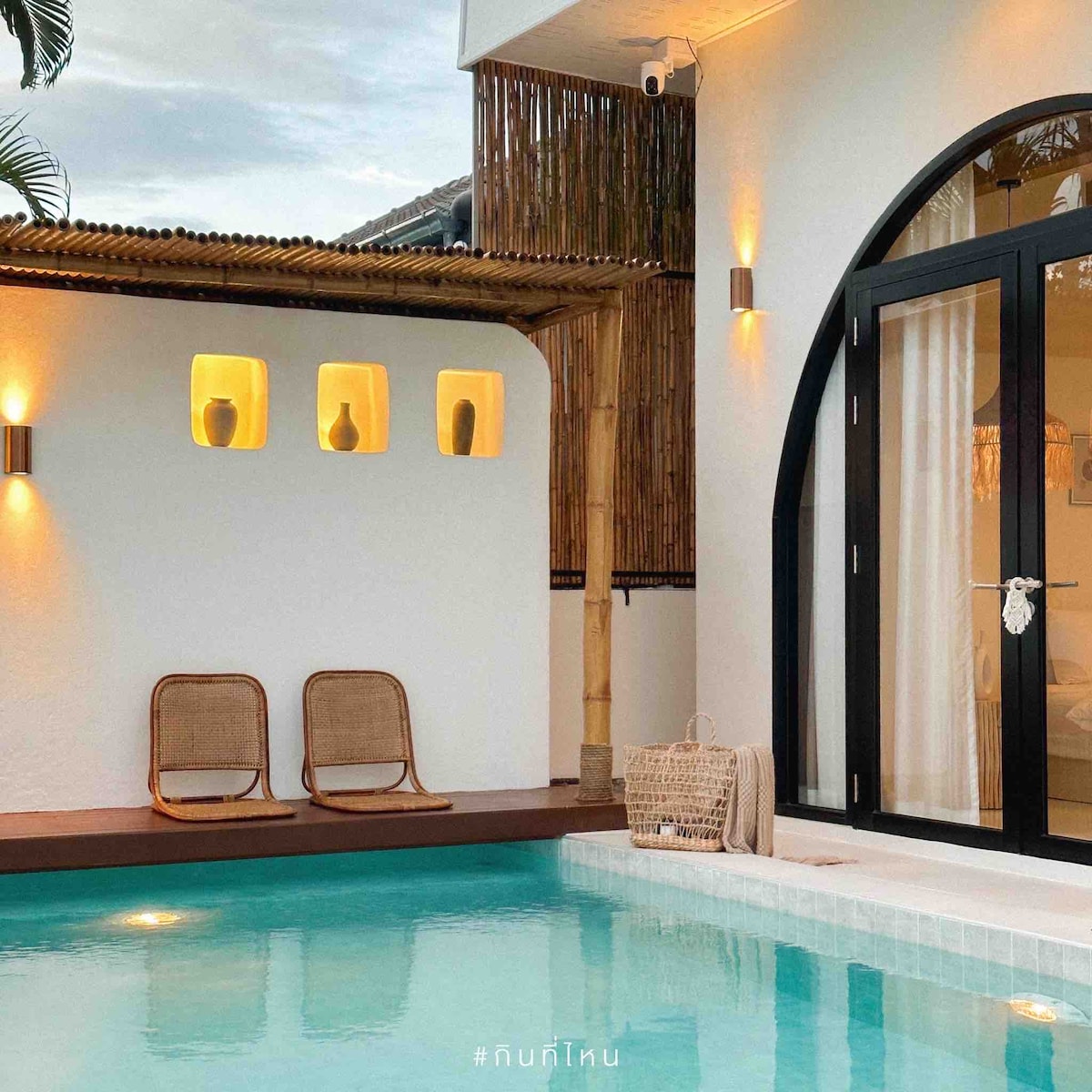An inviting outdoor area showcases a private swimming pool, complemented by two woven chairs positioned nearby. Soft lighting highlights the textured walls and decorative shelves, which display minimalist vases, enhancing the serene ambiance of the space.
