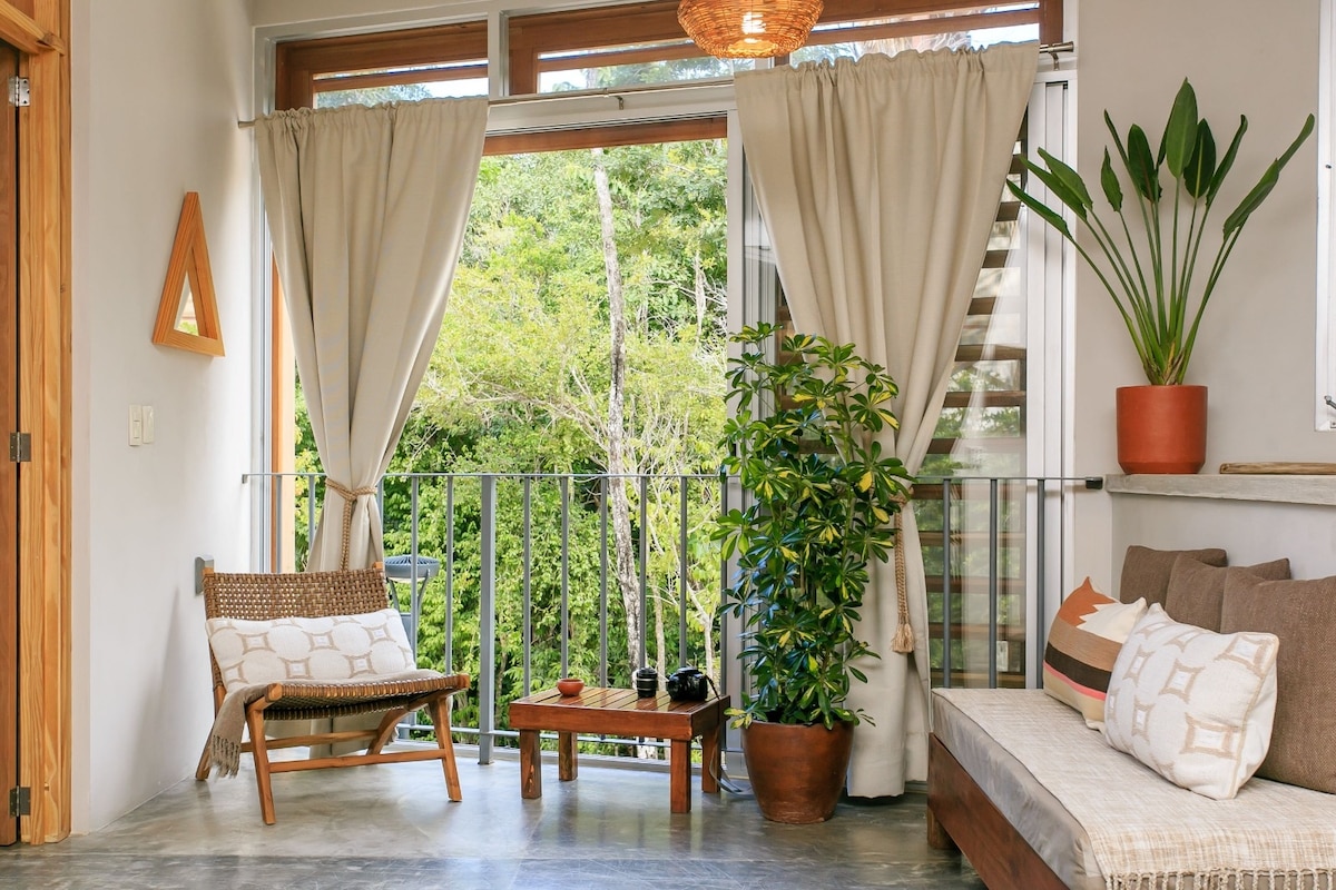 A cozy seating area features natural light streaming through large windows dressed with light curtains. A potted plant adds a touch of greenery beside a comfortable sofa and a wooden chair. The outdoor view includes lush foliage, enhancing a sense of relaxation.