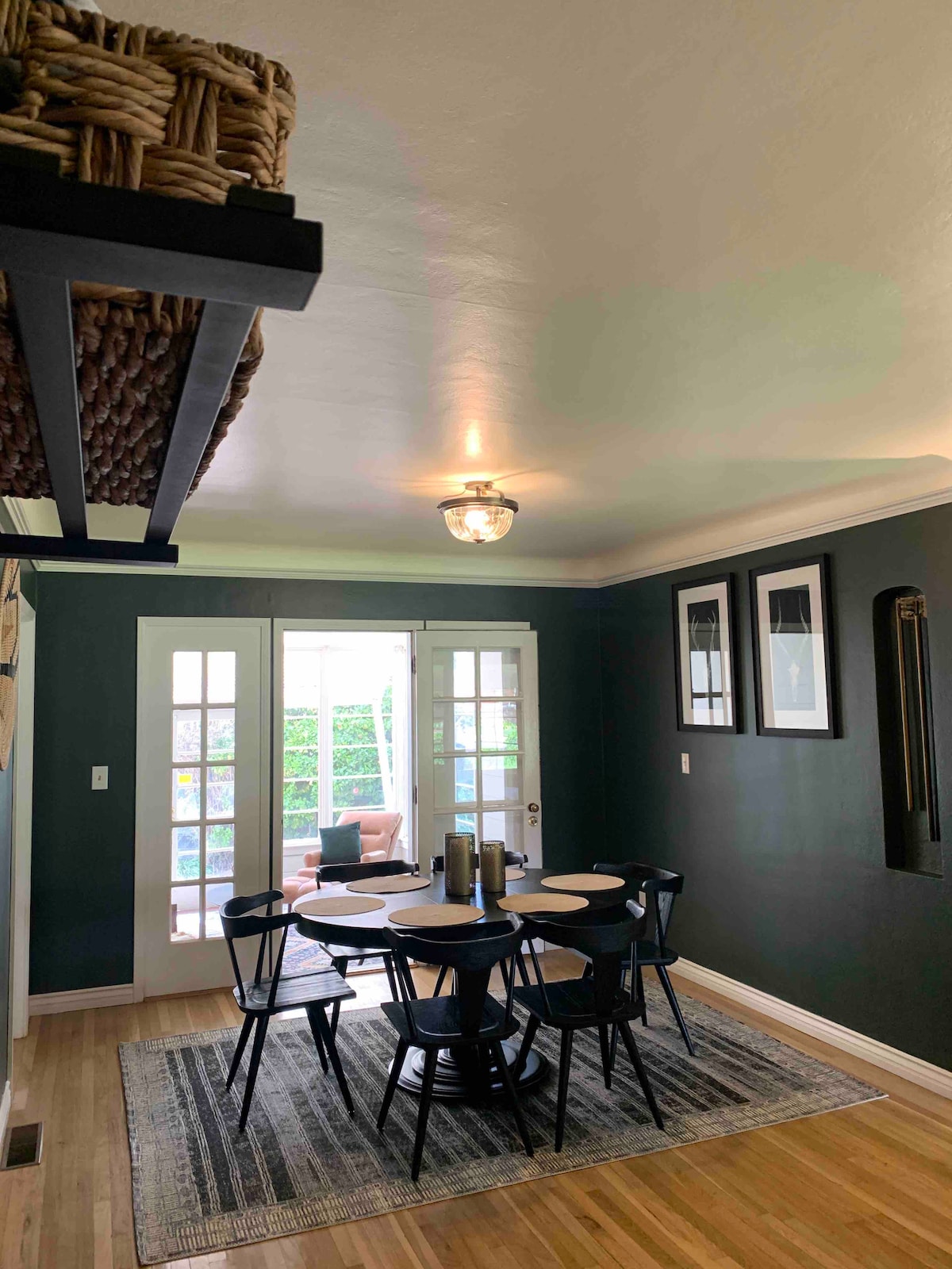 A dining area is presented with a round black table surrounded by six black chairs. Light-colored wooden flooring complements dark green walls, while sunlight filters through double French doors leading to an outdoor space. Decor includes framed artwork on the walls and a textured rug beneath the table.