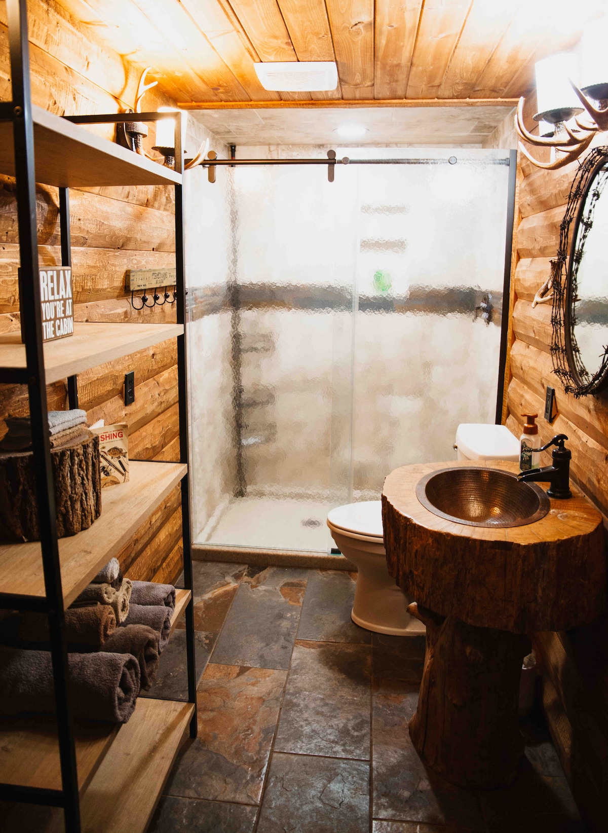 A rustic bathroom is highlighted by wooden accents and natural stone flooring. A standing shower with frosted glass doors is visible, along with a round wooden sink atop a sculpted base. Open shelving holds neatly rolled towels, enhancing the cabin's cozy aesthetic.
