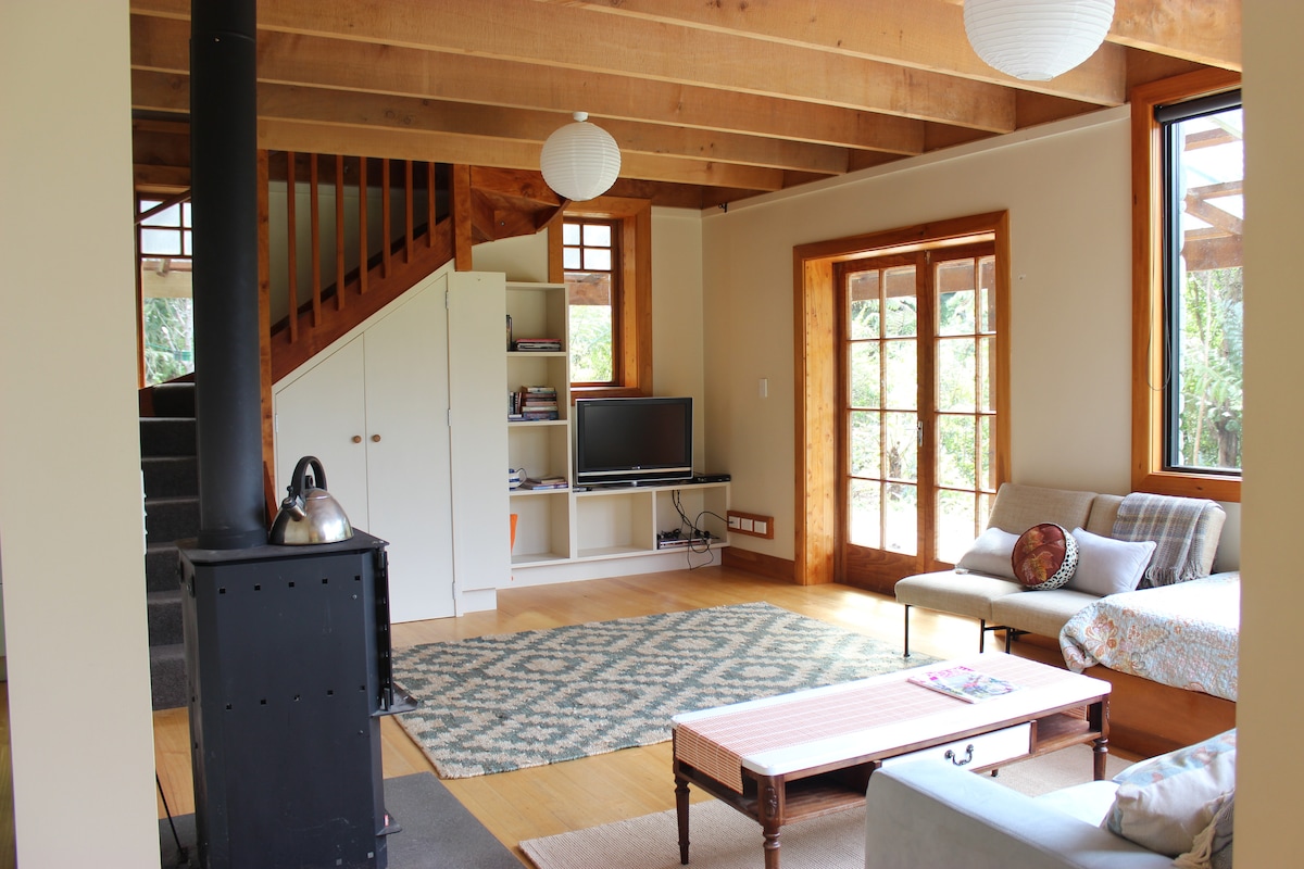 A spacious living area features wooden beams and large windows allowing natural light. A comfortable sofa and an antique coffee table are positioned on a patterned rug. A flat-screen television is mounted on the wall, with shelves nearby for books and decorative items.