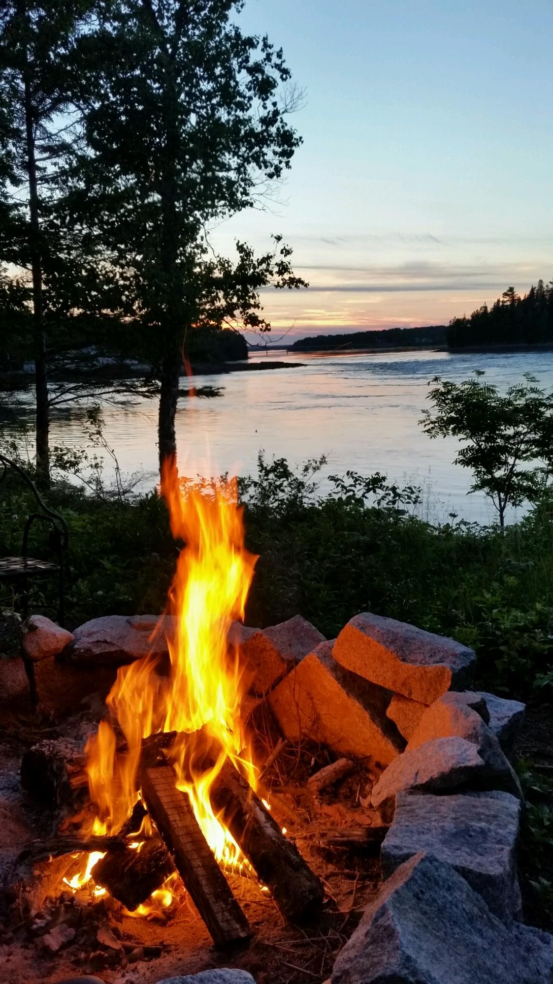 A fire pit surrounded by stone sits near a calm shoreline, with flames reaching upward against a backdrop of a tranquil river at sunset. Trees frame the scene, and the sky reflects a soft gradient of colors transitioning into evening.