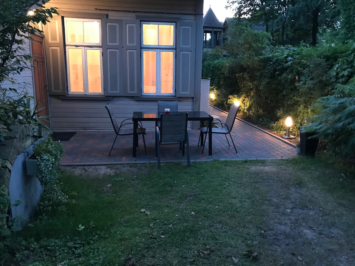 An outdoor seating area features a table with four chairs set on a paved patio, surrounded by shrubs and soft lighting. Beyond the patio, a grassy area extends, while large windows of the house reveal a warm glow from within.
