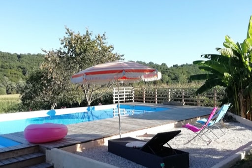 A shared swimming pool is surrounded by wooden decking and landscaping, featuring a colorful umbrella and lounge chairs. Lush green trees form a serene backdrop, emphasizing the tranquil environment. A circular float is visible in the water, adding a touch of leisure to the space.