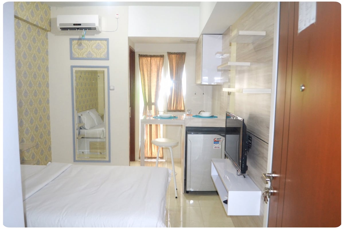 A compact living space features a light-colored bed with crisp linens. A small dining area with two stools is positioned near the kitchenette, which includes a mini fridge. Floor-to-ceiling windows allow natural light to fill the room, with sheer curtains softly draping.