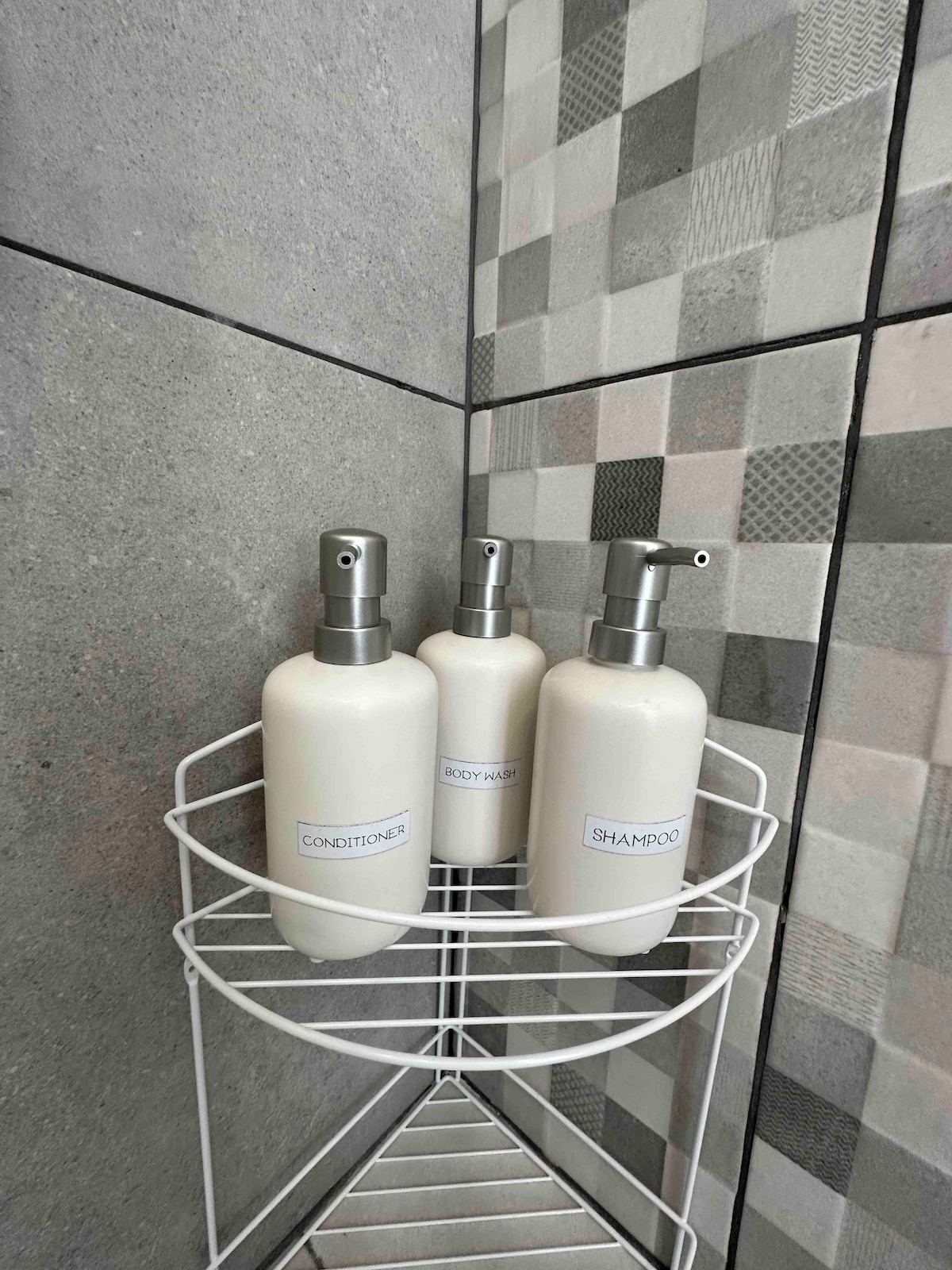 Three white pump bottles labeled 'Conditioner,' 'Shampoo,' and 'Body Wash' are displayed on a metal shower caddy. The wall features a modern tile design with a combination of gray and patterned tiles.