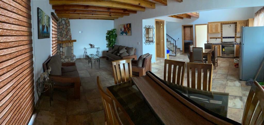 A spacious living area features wooden beams overhead and a stone accent wall. Comfortable seating options include a sofa and armchairs, complemented by a small coffee table. The open layout leads to a dining area, which includes a wooden table and chairs, along with a kitchen visible in the background.