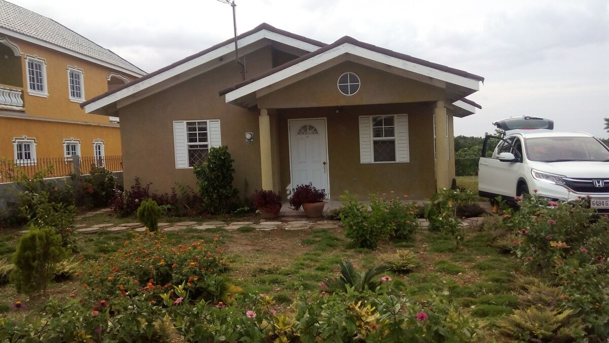 The front of the house is presented, featuring a welcoming entrance with a white door and distinctive round window. Colorful flowers and greenery are arranged in the yard, with stone pathways leading to the door. A vehicle is parked nearby.