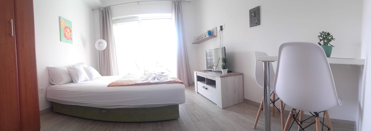 A studio apartment is showcased with a spacious bed dressed in white linens, complemented by a colorful throw. A television unit sits against the wall, and a small dining table with two chairs is positioned nearby. Natural light floods the room through a large window with sheer curtains.