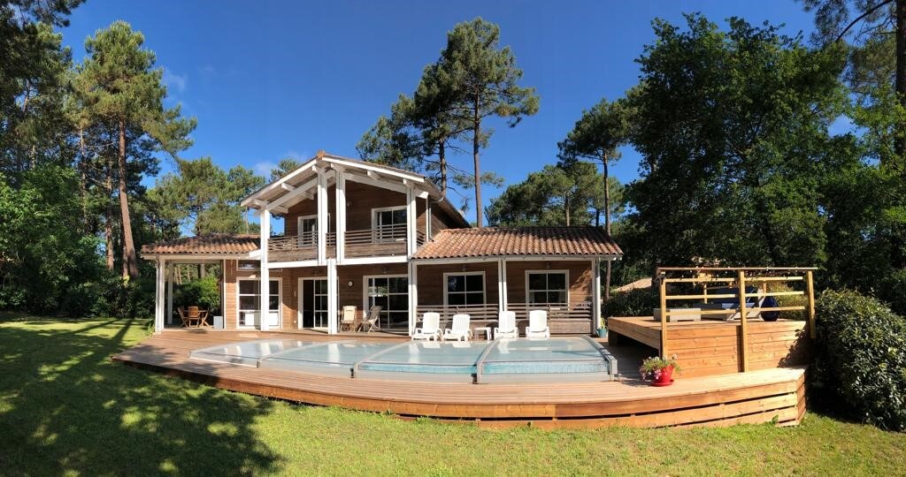 The villa is surrounded by lush greenery and features a spacious wooden deck with a private heated pool. Sun loungers are positioned nearby, inviting relaxation. Floor-to-ceiling windows enhance natural light, and a balcony is visible on the upper level, contributing to the property’s inviting appeal.