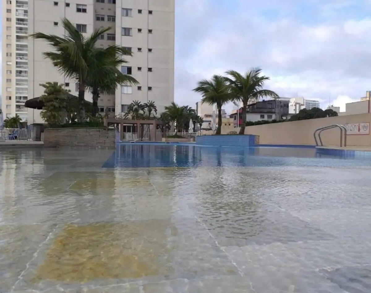 A spacious pool area features calm waters surrounded by palm trees. The pool is divided into two sections, with a shallow area and a deeper section visible. Sun loungers and shaded seating are positioned alongside, inviting relaxation.