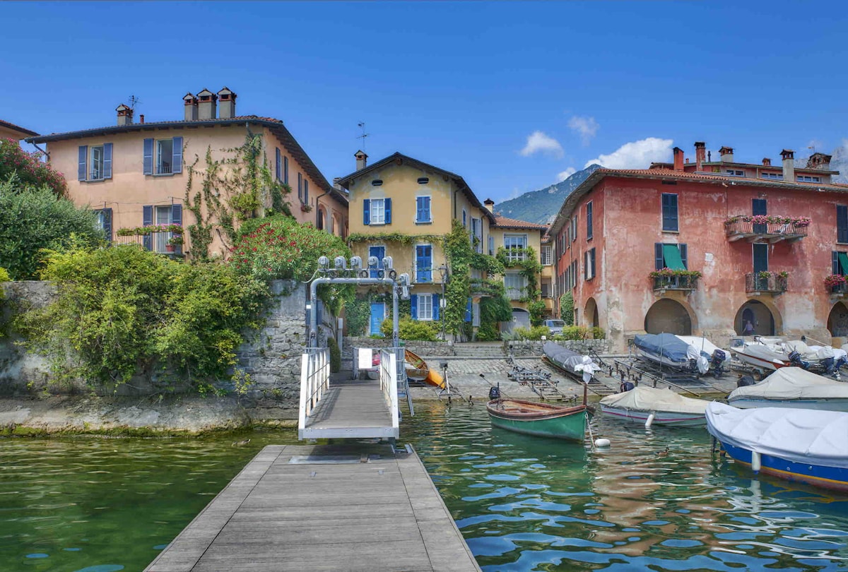 A wooden dock extends over calm waters, providing access to various small boats. Colorful buildings with balconies and greenery are visible in the background, reflecting a charming lakeside environment. The surrounding area shows a mix of architectural styles, set against a clear blue sky.
