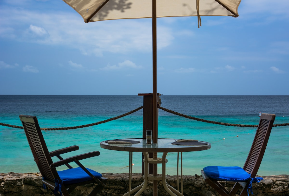 A table and two chairs are positioned under a large umbrella, overlooking a calm sea. The turquoise water is visible in the background, creating a serene setting for relaxation. Soft clouds are observed in the sky, enhancing the tranquil atmosphere.