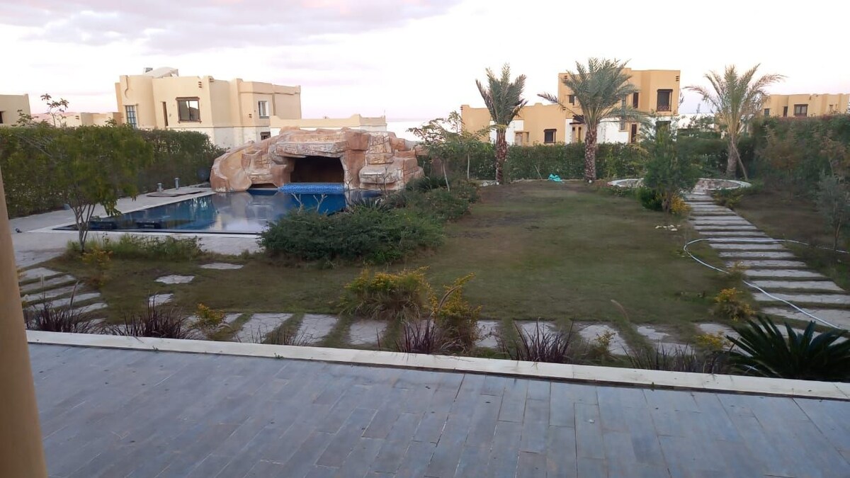 A spacious outdoor area features a landscaped garden with palm trees and a swimming pool shaped like a natural oasis. Stone pathways lead through the garden, connecting different areas of lush greenery and decorative plants.