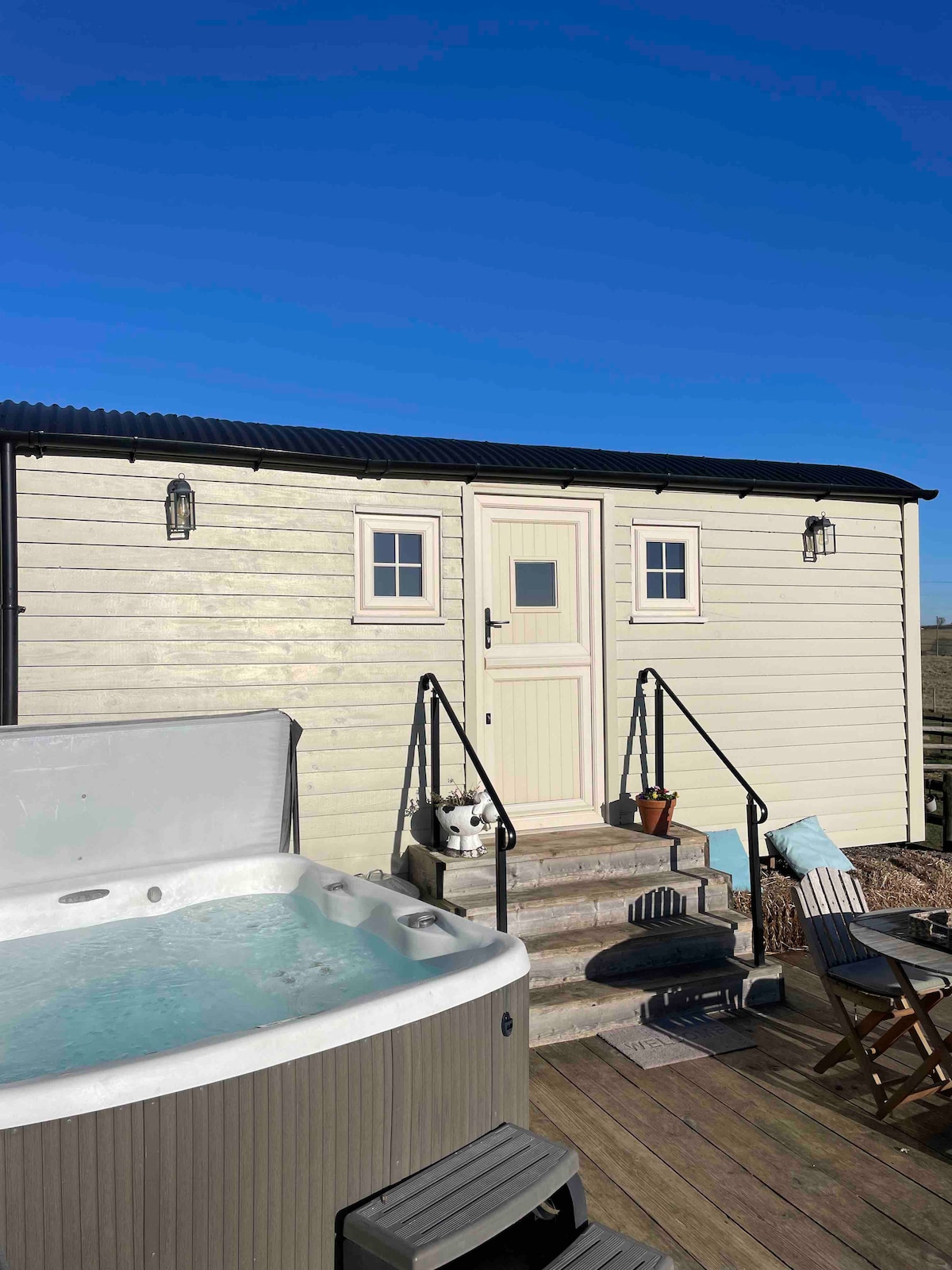 A luxury shepherd's hut is featured, showcasing a light-colored exterior with two small windows flanking the entrance door. Steps lead up to the entry, and a hot tub is positioned nearby, inviting relaxation with clear water glistening in the sunlight.