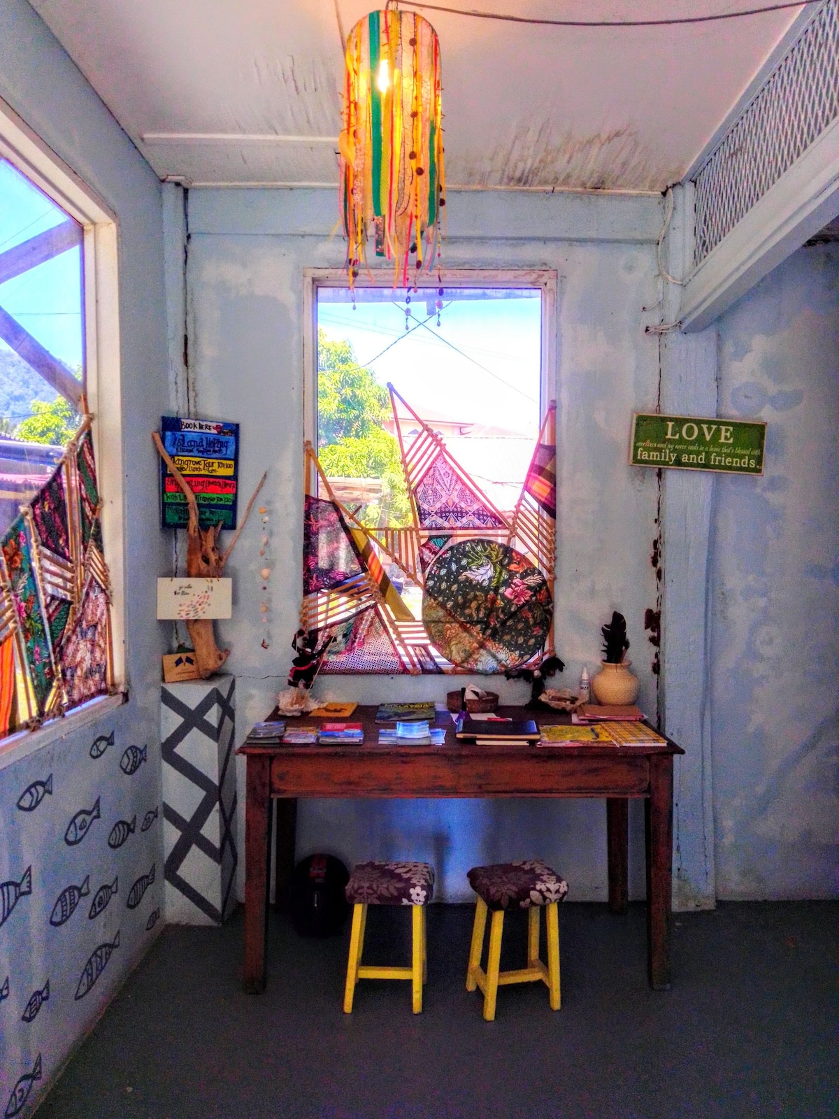 A small entryway features a wooden table adorned with books and decorative items. Colorful, patterned curtains frame the windows, allowing natural light to brighten the space. A unique light fixture hangs above, while two small stools are positioned nearby.