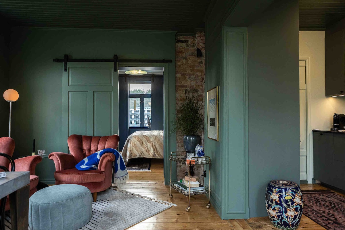 A cozy living area displays a plush armchair and a soft ottoman, complemented by elegant decor. A glimpse of a spacious bedroom can be seen through the doorway, with a warm-toned bedspread and subtle lighting. Exposed brick adds texture to the green walls.