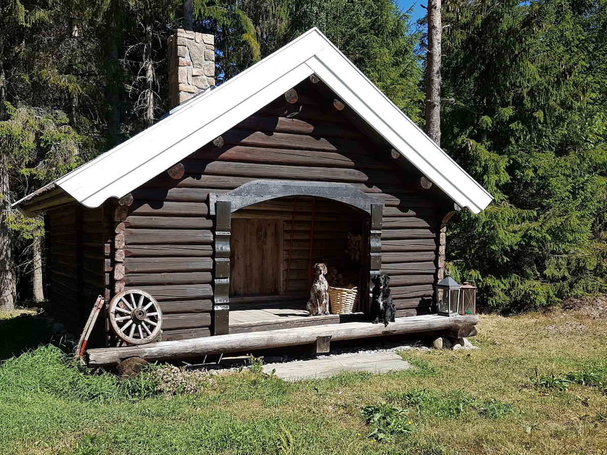 A traditional wooden cabin is depicted, featuring a sloping roof and an inviting porch. Two dogs are positioned on the porch, while a wooden wheel and basket are placed nearby, surrounded by lush greenery and towering trees in the background.