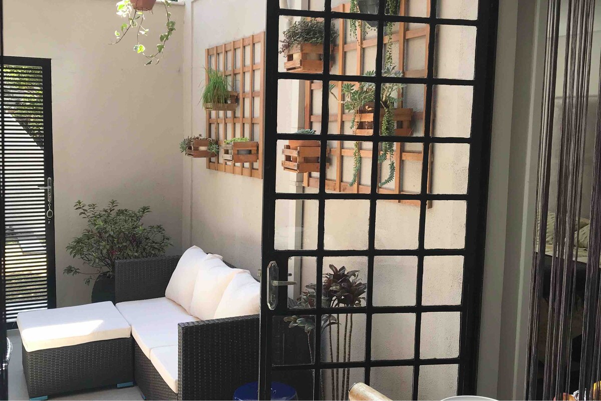 An outdoor seating area is displayed, featuring a dark wicker couch adorned with light cushions. Vertical planters with greenery are attached to the wall, contributing to a tranquil ambiance. The entrance is framed by black grid-patterned doors that separate the space from the interior.
