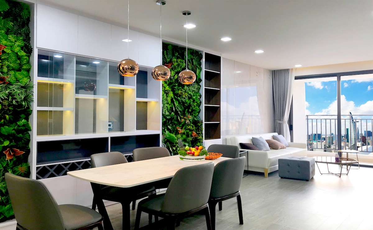 The spacious dining area features a light wooden table surrounded by six chairs, complemented by a decorative wall adorned with greenery. Large windows provide a view of the city skyline and allow natural light to illuminate the space, enhancing the contemporary design.