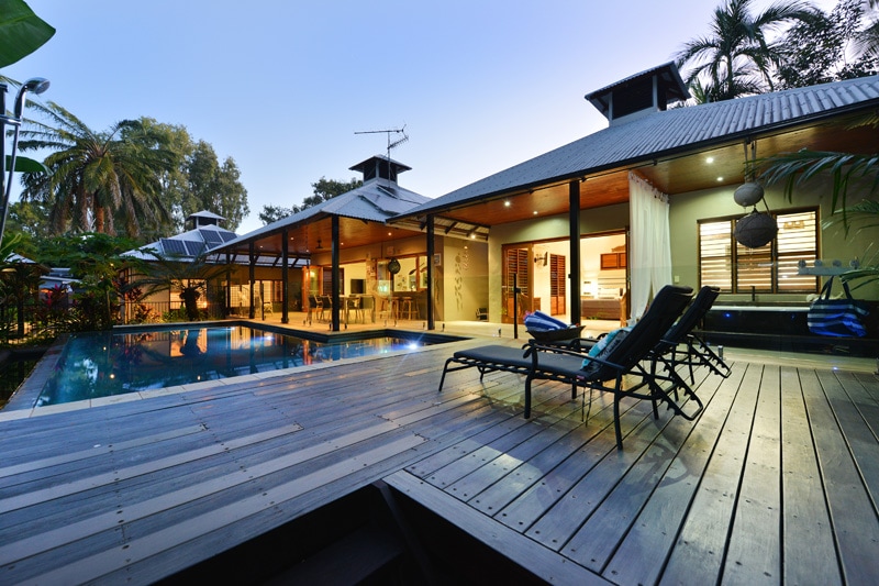 The outdoor area features a wooden deck surrounding a swimming pool, framed by lush tropical plants. Lounge chairs are positioned neatly along the side, inviting relaxation. The pavilion-style home in the background has multiple large windows, allowing for a view of the adjoining indoor spaces.