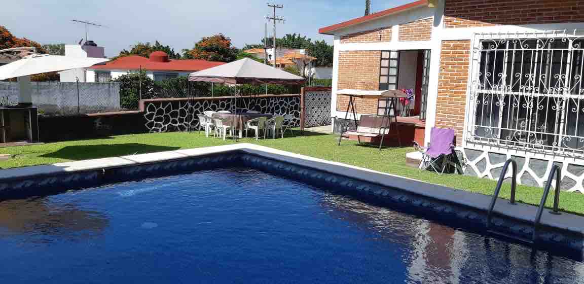 A private pool glistens under the sun, surrounded by a manicured lawn. A seating area with tables and chairs is shaded by a canopy nearby. The rustic brick house features large windows, allowing natural light to fill the interior spaces.