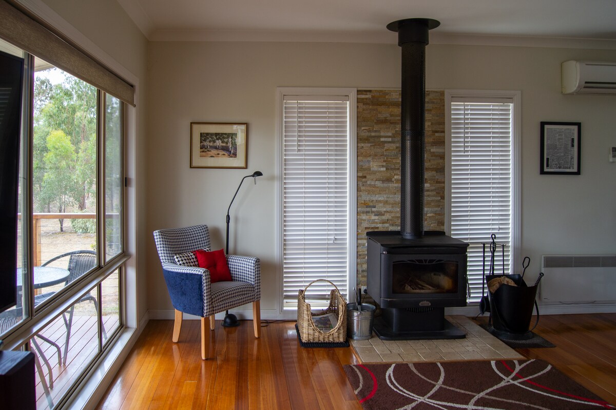 A cozy living area features a wood heater surrounded by modern furnishings. A comfortable chair with a blue cushion is positioned nearby. Large windows allow natural light to brighten the space, and wooden flooring adds warmth. A soft rug is placed on the floor.