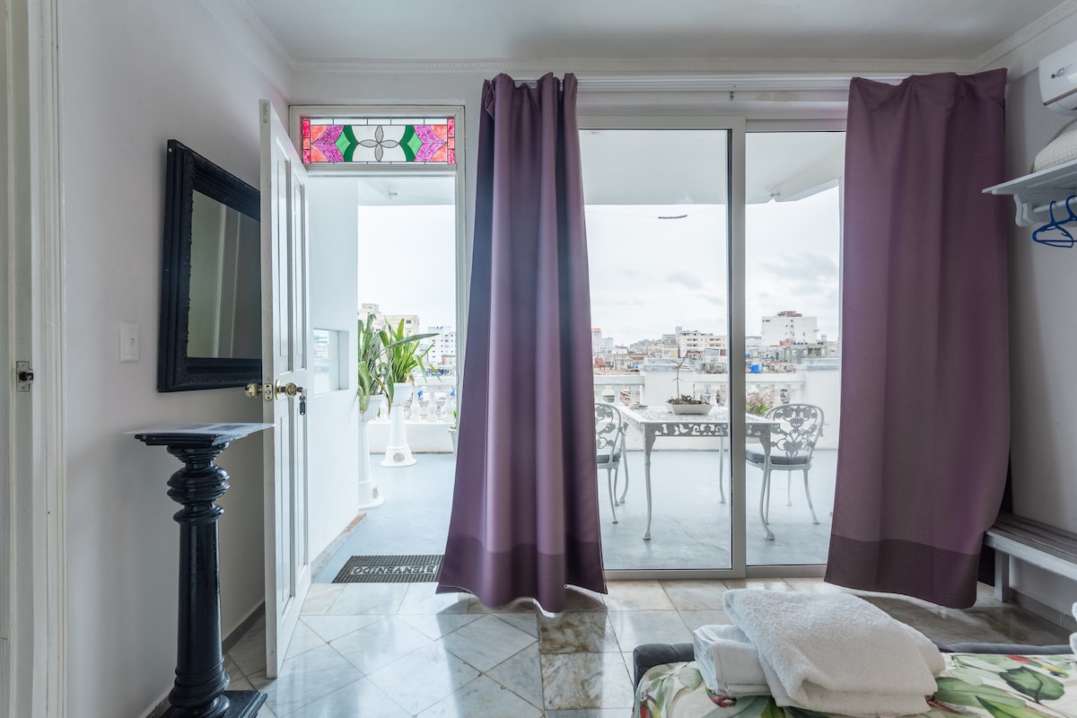 Sheer curtains frame a large sliding glass door that opens to a balcony with a view of the city and sea. A decorative stained glass panel is visible above the door, and a small console table adds functional charm to the space.