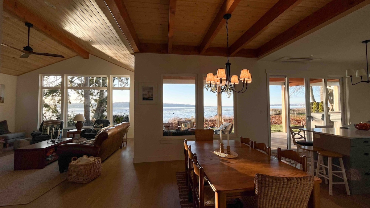 An open-concept living area features high ceilings with exposed beams and large windows showcasing ocean views. A spacious dining table is surrounded by wooden chairs, while a light fixture hangs overhead. Cozy seating is arranged nearby, creating a welcoming gathering space.