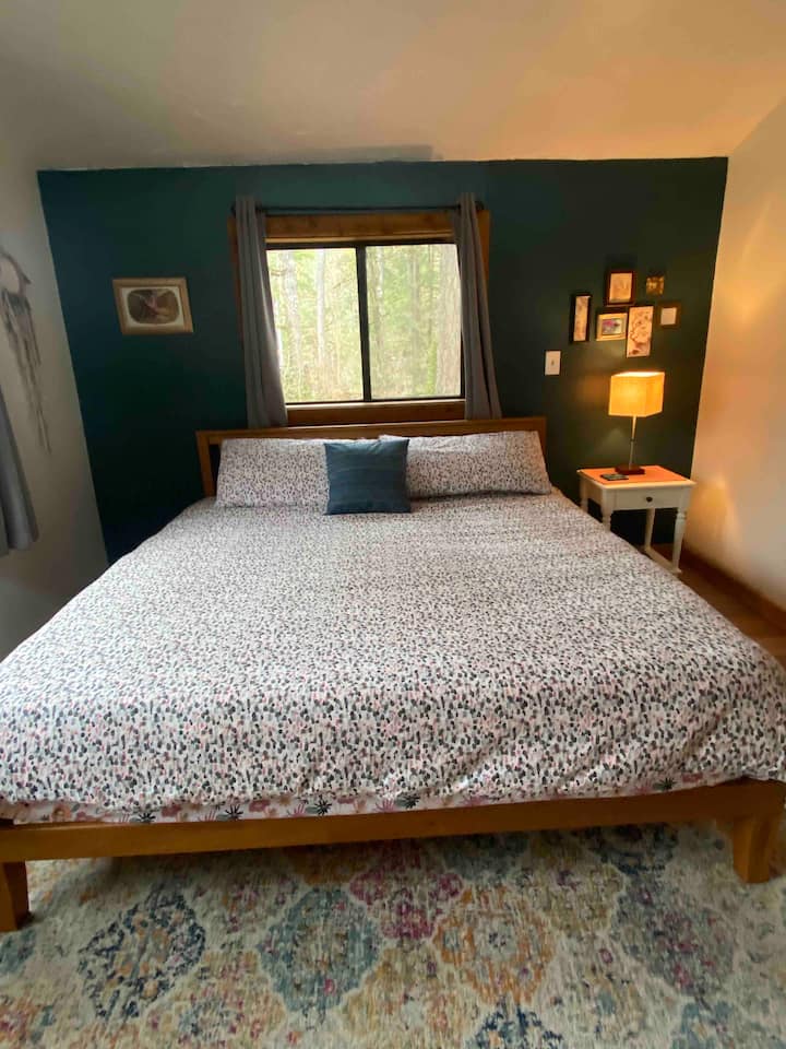 King bedroom with new king mattress, new bed frame, and cozy greenish blue paint. Next picture shows wall of windows looking out to forest.