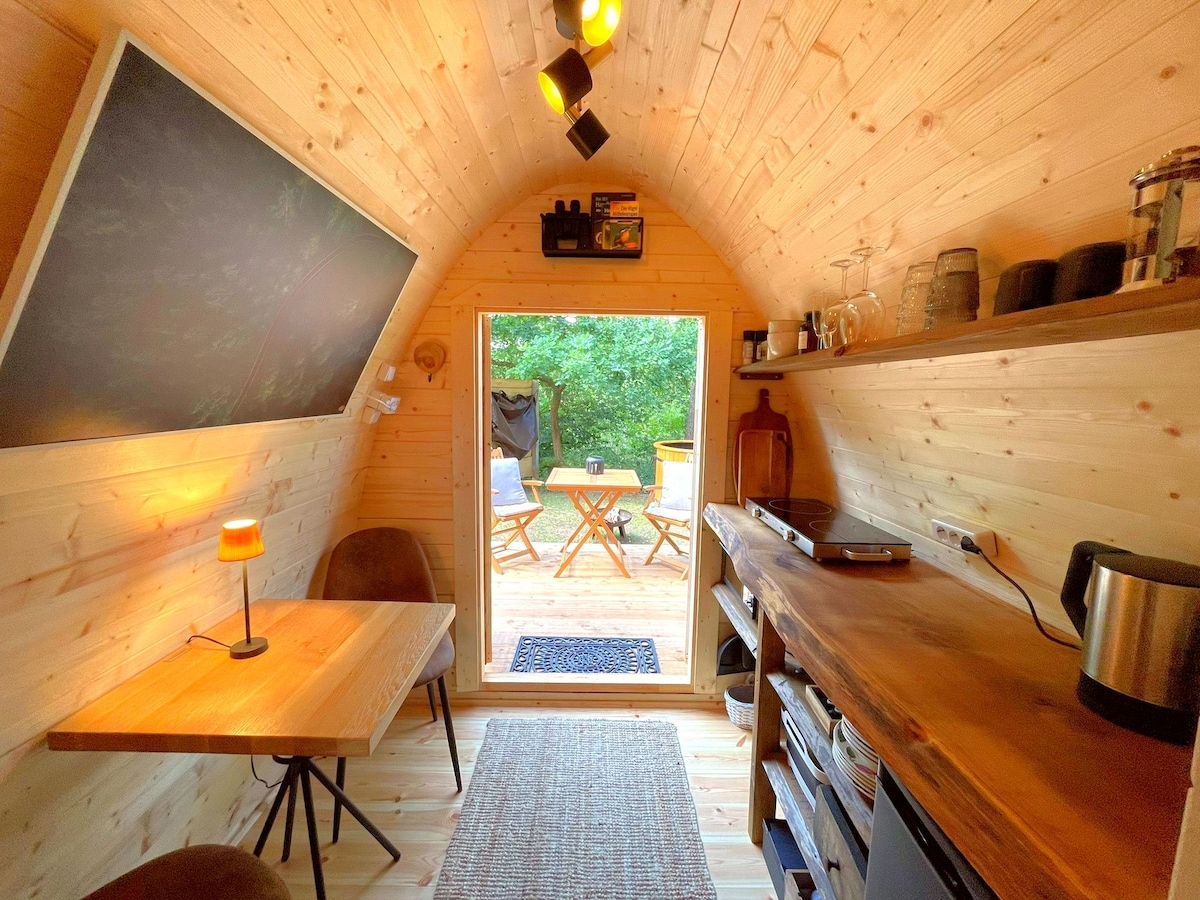 The cozy interior of a glamping pod features warm wooden walls, a small dining table, and seating options. A kitchenette is present, equipped with basic appliances. A door opens to a deck area, showcasing a natural view of the surrounding woodland.