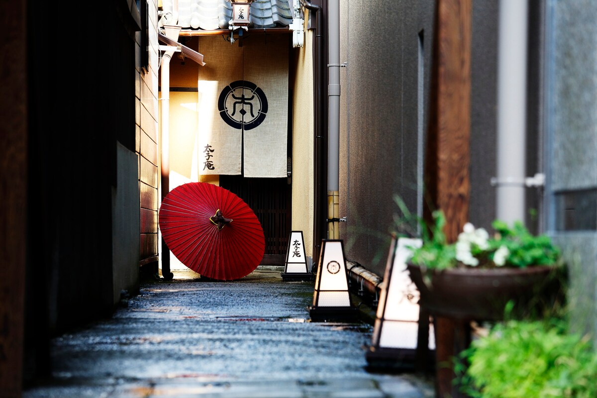 A narrow alleyway is lined with traditional lanterns and a vibrant red umbrella resting against a wall. Soft lighting creates a serene ambiance as the path leads to a subtle entrance adorned with Japanese characters, inviting exploration of the surrounding area.