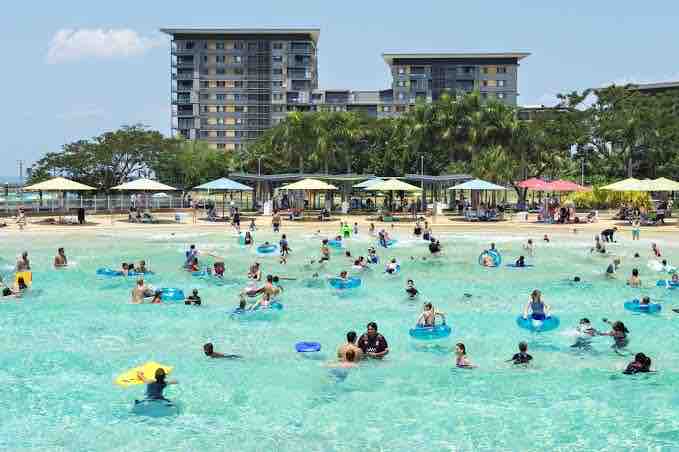 A vibrant scene captures visitors enjoying the water at a beach area, surrounded by lush greenery and colorful umbrellas. The clear water is filled with people using various inflatable devices, while sandy areas provide space for relaxation. Modern buildings are visible in the background.