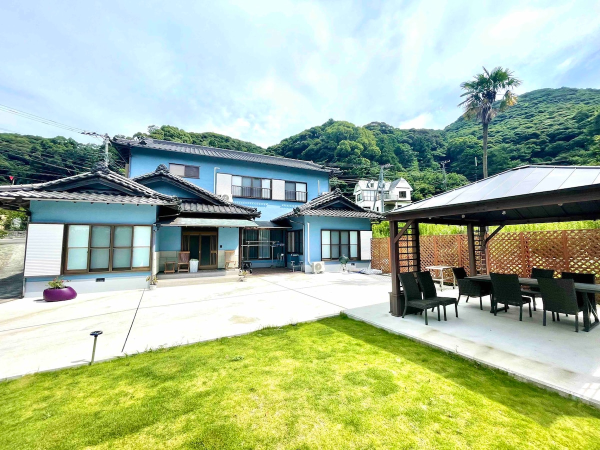 A large traditional Japanese house is positioned amidst lush greenery. The two-story structure features a welcoming outdoor space with a spacious patio and a gazebo equipped with a dining area. The well-maintained lawn adds to the serene environment surrounding the property.