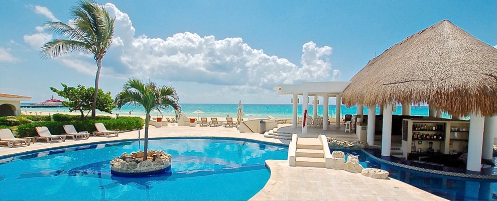 A spacious pool area features an inviting blue water pool surrounded by lounge chairs and palm trees. A thatched-roof bar is visible, providing a shaded area. The beautiful Caribbean Sea serves as a stunning backdrop under a bright sky dotted with clouds.