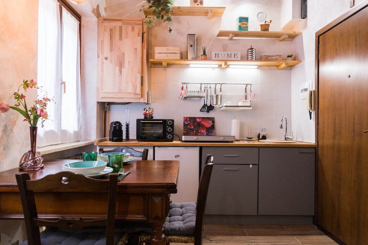 The open kitchen area features wooden shelves displaying kitchen essentials alongside modern appliances, including a microwave and electric oven. A dining table with two place settings is positioned next to the window, allowing natural light to illuminate the space.