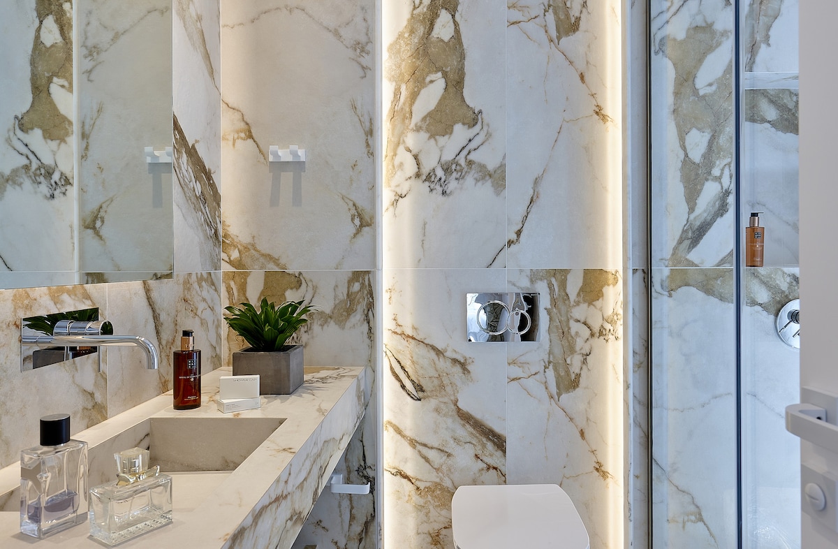 The bathroom features elegant marble walls with striking gold veining. A modern sink is situated on a spacious countertop, while a glass shower enclosure is visible, showcasing sleek fixtures. Decorative elements include a small potted plant and neatly arranged toiletries.