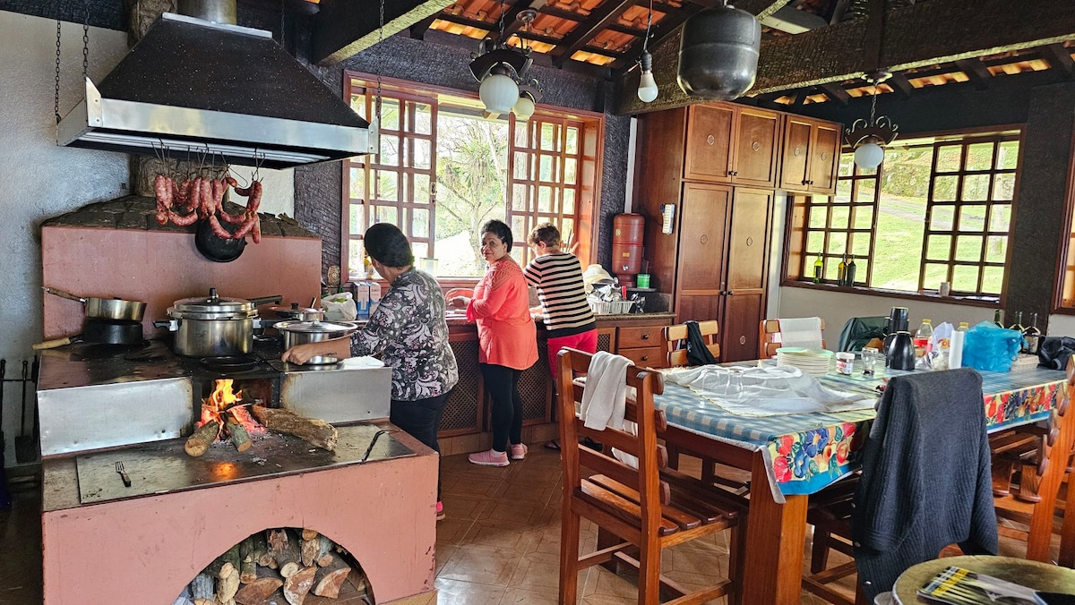 A functional kitchen space is depicted, showcasing a large cooking area with a stove and an open flame. Wooden cabinets and dining table are visible, while three individuals are engaged in meal preparation, highlighted by natural light streaming through the windows.