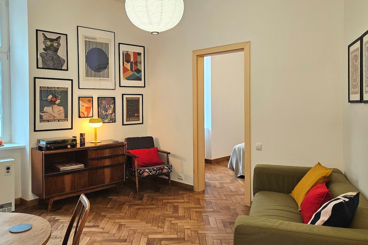 A cozy living area is presented, featuring a green sofa adorned with red and yellow cushions. Artwork in various frames is displayed on the walls, while a wooden sideboard holds a record player. Soft lighting casts a warm glow over the space.