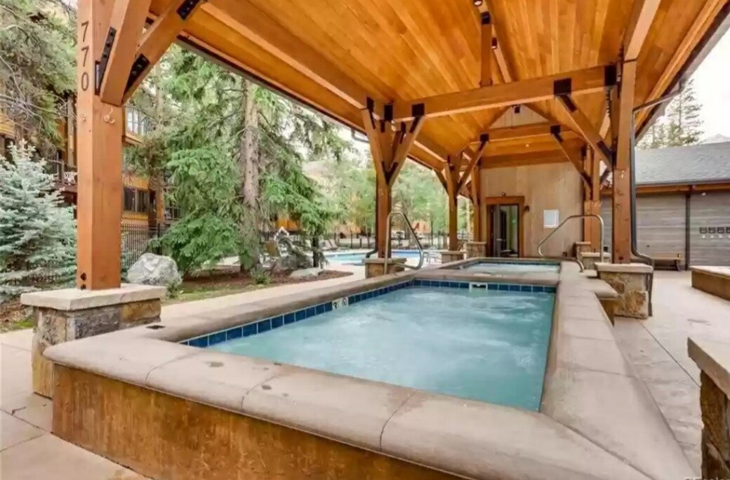 An outdoor hot tub is framed by wooden beams and a sloped ceiling, surrounded by trees and landscaped gardens. The tub features a stone perimeter and inviting water, with a view of the nearby pool area in the background.
