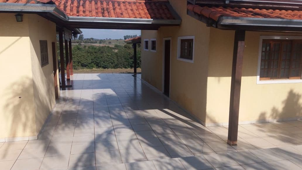 An outdoor patio space is presented, featuring a smooth tiled surface that extends adjacent to the building. The area is bordered by walls and overhanging roof sections, offering shaded spots. A lush green landscape is visible in the background, complementing the serene outdoor setting.