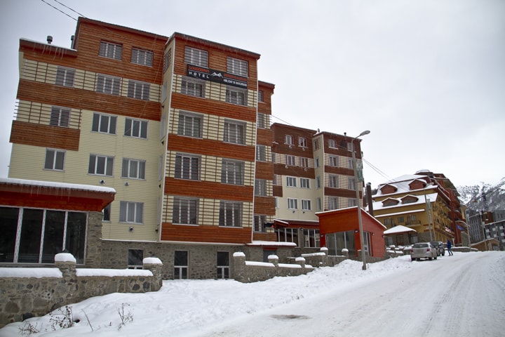 A multi-story building is presented with a combination of wood and stone finishes. Snow covers the ground, while the street is lined with light traffic. Large windows can be seen on each floor, allowing natural light to enter the interior spaces.
