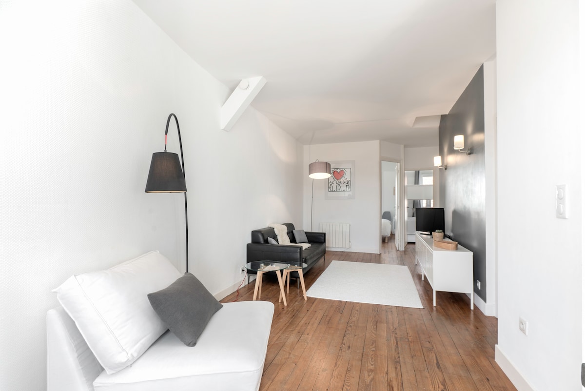 A bright living area features light-colored walls and hardwood flooring. A white sofa with accent pillows sits beside a black leather sofa. A minimalist lamp and two wall sconces provide soft lighting. A neutral area rug defines the space, offering a cozy atmosphere.