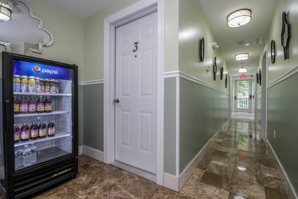 A well-lit hallway features smooth marble flooring and soft green walls. A small refrigerator stocked with drinks is positioned next to a door marked with the number three. Decorative mirrors and wall sconces add to the inviting atmosphere.